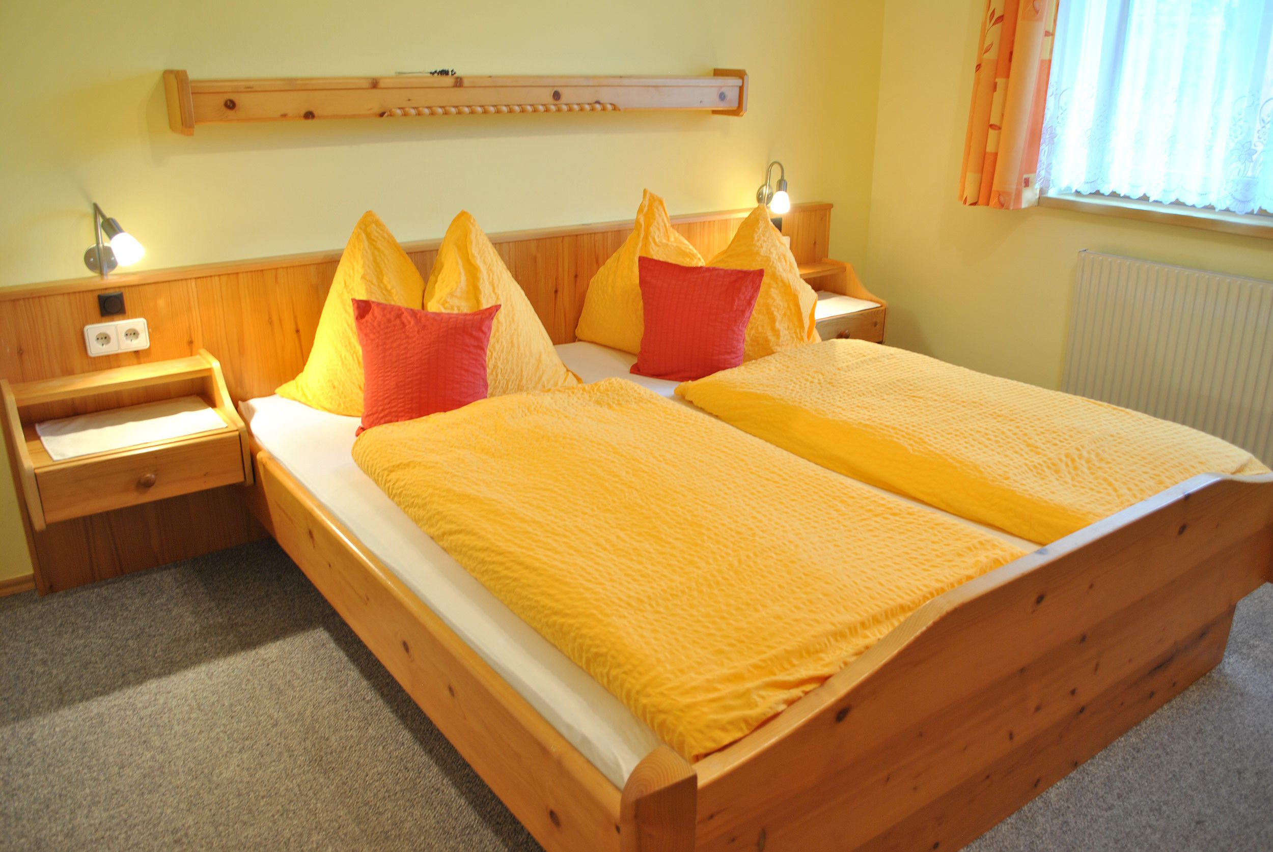 Una cama doble con estructura de madera y colcha amarilla, dos mesitas de noche con lámparas, un estante de madera sobre la cama y una ventana con cortinas.