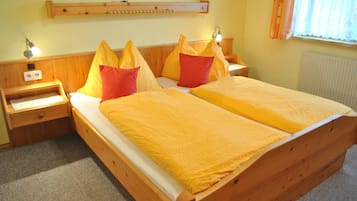 Una cama doble con estructura de madera y colcha amarilla, dos mesitas de noche con lámparas, un estante de madera sobre la cama y una ventana con cortinas.
