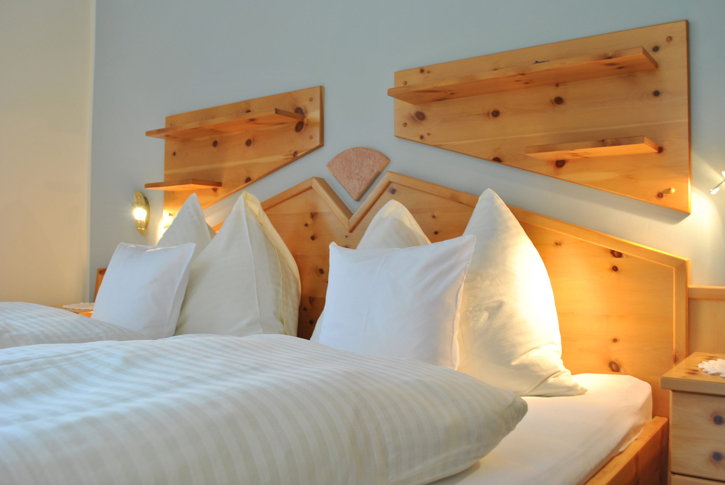 Una cama con ropa de cama y almohadas blancas, un cabecero de madera con diseño geométrico y una mesita de noche con una lámpara.