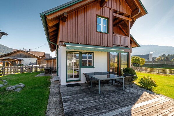 Een houten huis met twee verdiepingen, met een balkon, een terras met tafel en stoelen, en een uitzicht op de bergen op de achtergrond.