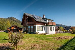 Een groot houten huis met een schuin dak, een apart wit gebouw met groene accenten, en een omheind erf.