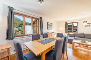 Een moderne woonkamer met een houten eettafel, grijs gestoffeerde stoelen, een grijze bank en uitzicht op een berg door grote ramen.