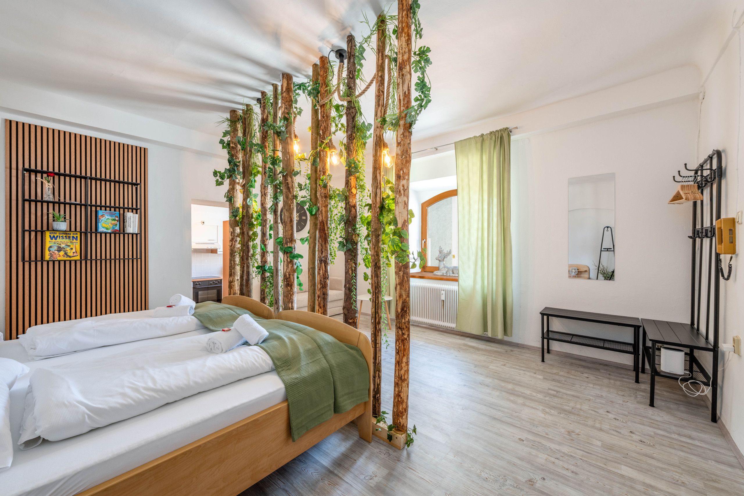 Una camera d'albergo con un letto, una testiera in legno, una mensola a parete con libri e una pianta, una panca e vista sull'area cucina.