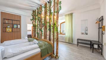 Una camera d'albergo con un letto, una testiera in legno, una mensola a parete con libri e una pianta, una panca e vista sull'area cucina.