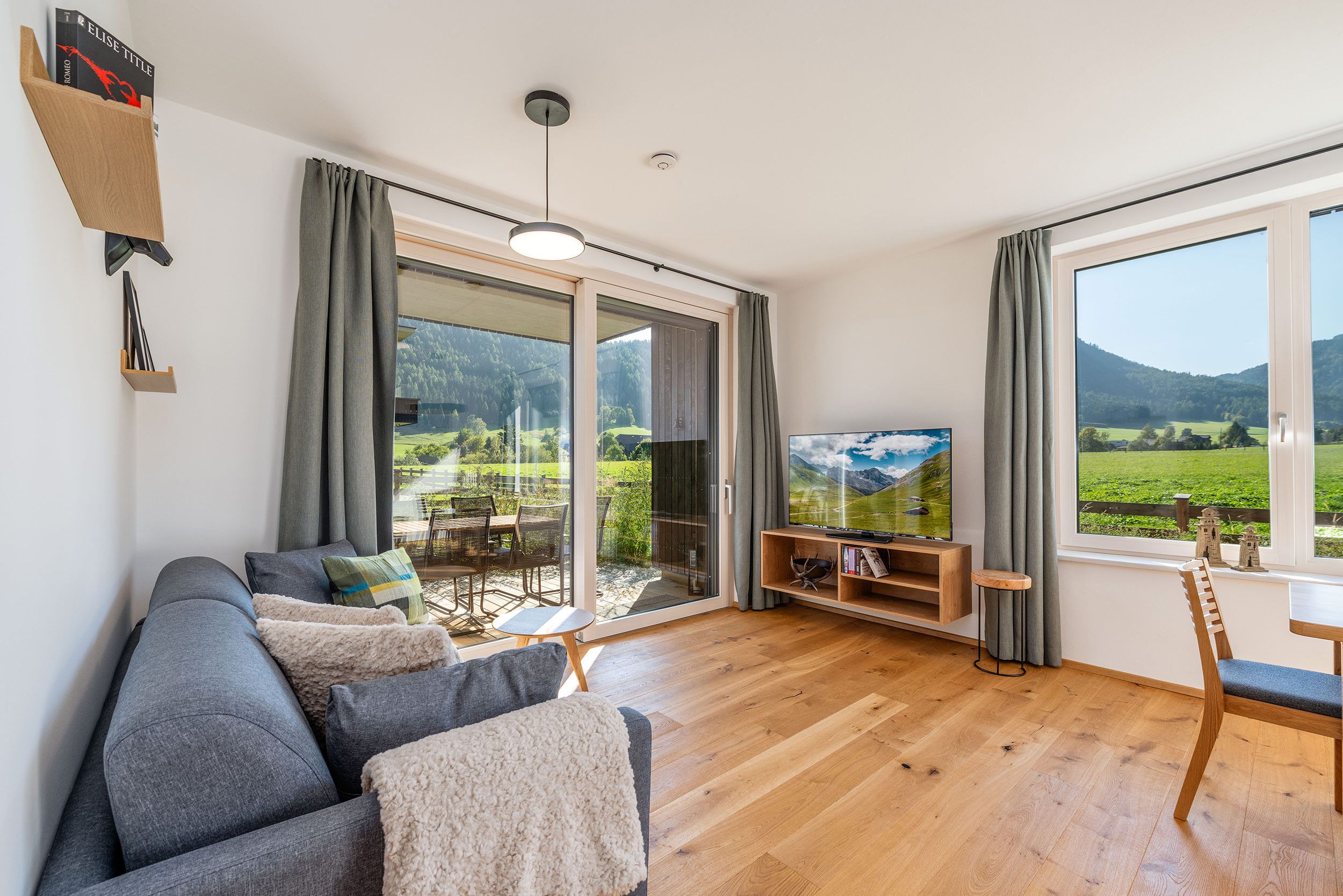 Un salon moderne avec un canapé, un téléviseur et une vue sur un paysage verdoyant grâce à de grandes fenêtres.