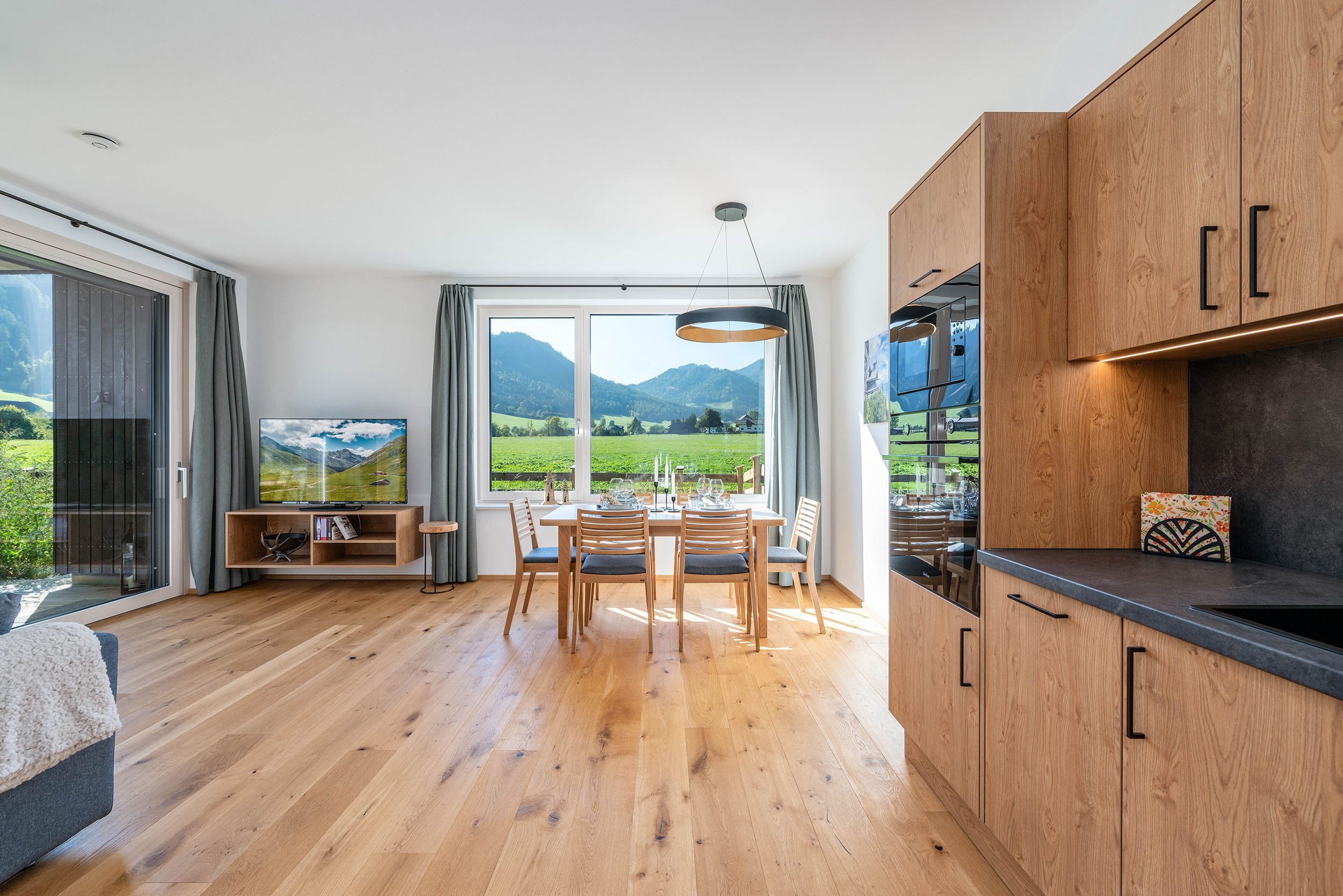 A modern kitchen with wooden cabinets, a dining area with a table and chairs, and a large window offering a view of a green landscape.