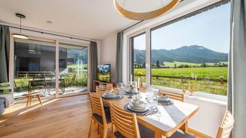 Een eetruimte met een tafel gedekt voor vier personen, grote ramen met uitzicht op een groen landschap en bergen, en een modern interieur.