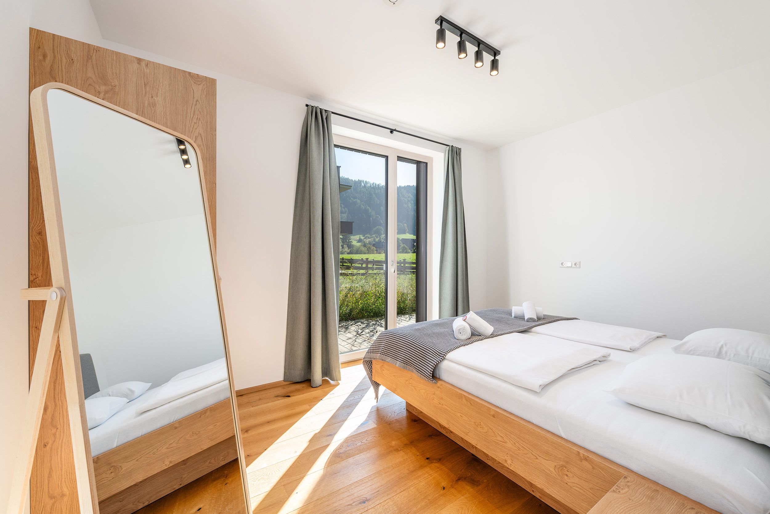 A modern bedroom with a wooden bed frame, a large mirror, and a view of a green landscape through a sliding door.