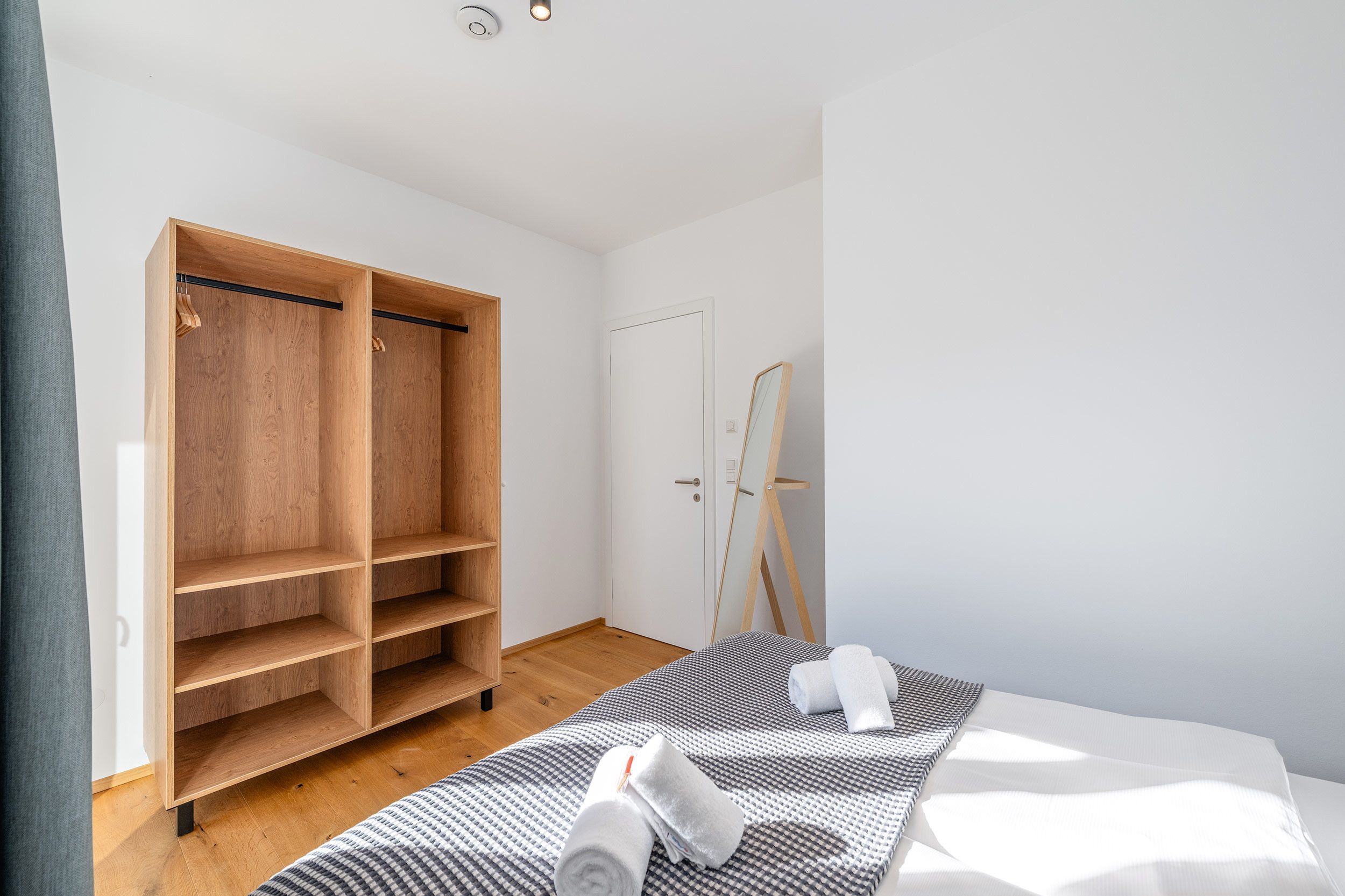 Een slaapkamer met een houten garderobe, een bed met een geruit sprei en een spiegel die tegen de muur staat.