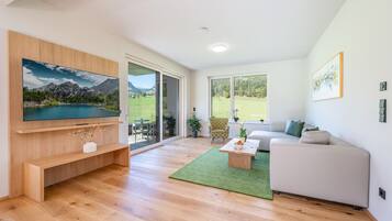 A modern living room with a large flat-screen TV, a sofa, a coffee table, and a view of a green landscape through the glass door.