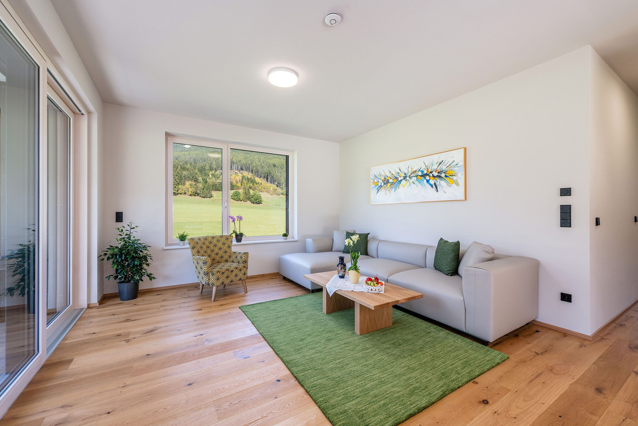 A modern living room with a large window, a sofa, a patterned armchair, a coffee table with a vase and fruit, and a green rug.