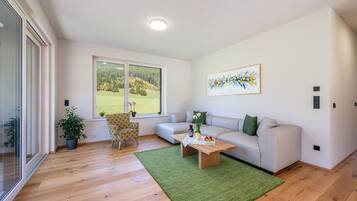A modern living room with a large window, a sofa, a patterned armchair, a coffee table with a vase and fruit, and a green rug.