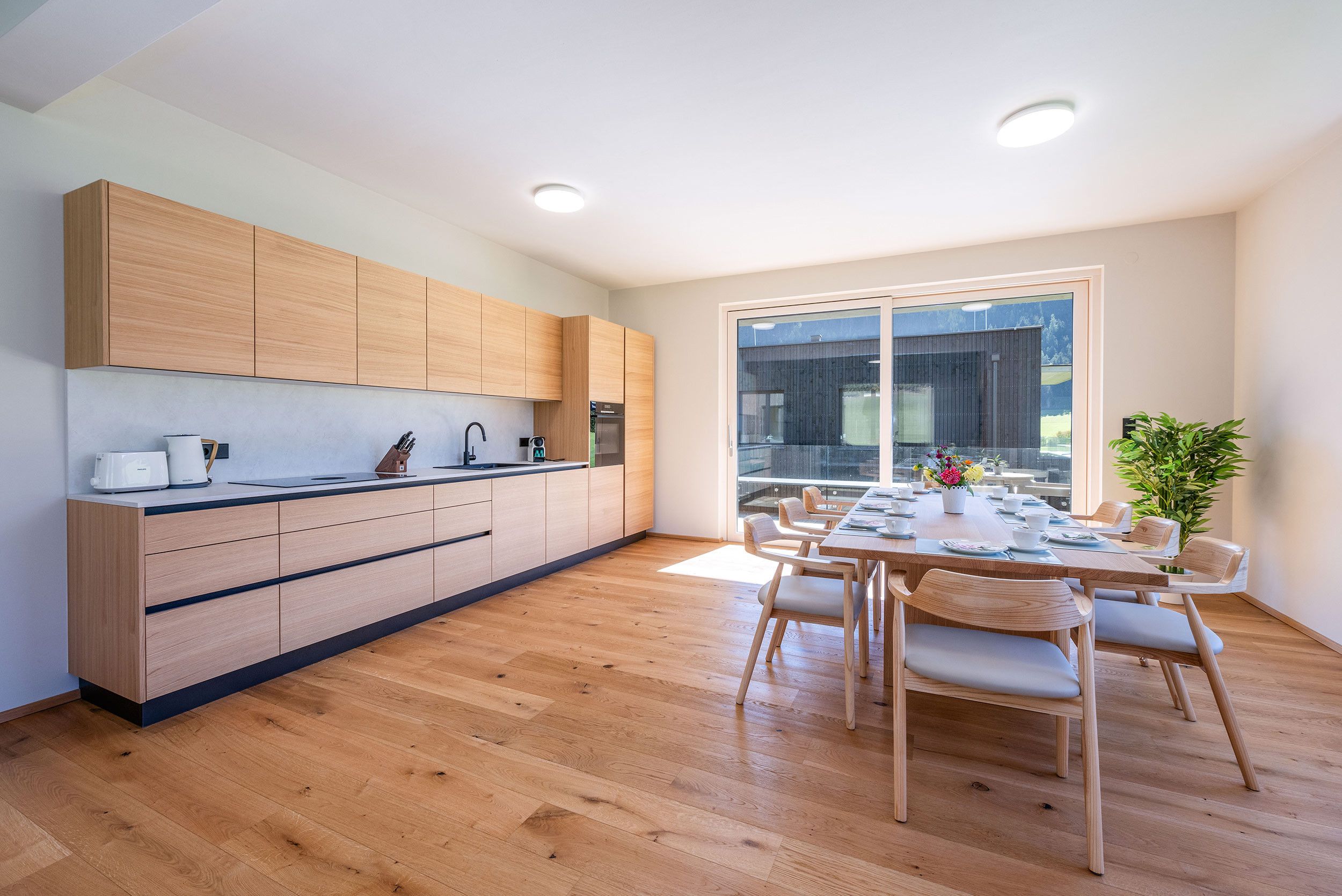 A modern kitchen with wooden cabinets, a dining area with a table and chairs, and a large window offering a view.