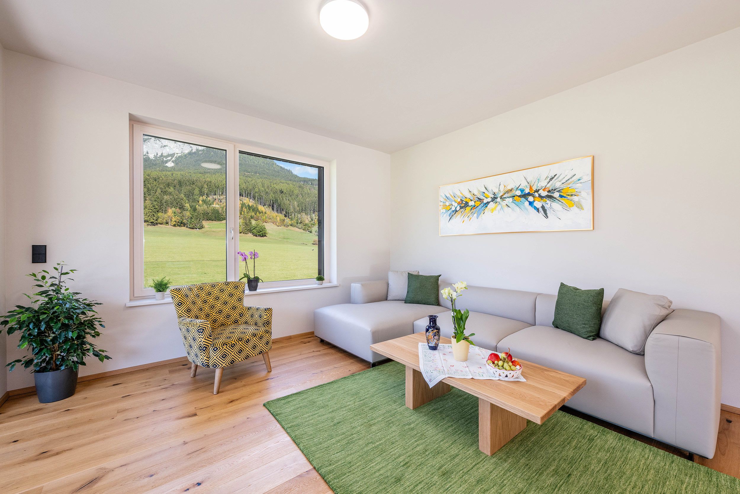 A modern living room with a large window, a sofa, a patterned armchair, a coffee table, and a painting on the wall.