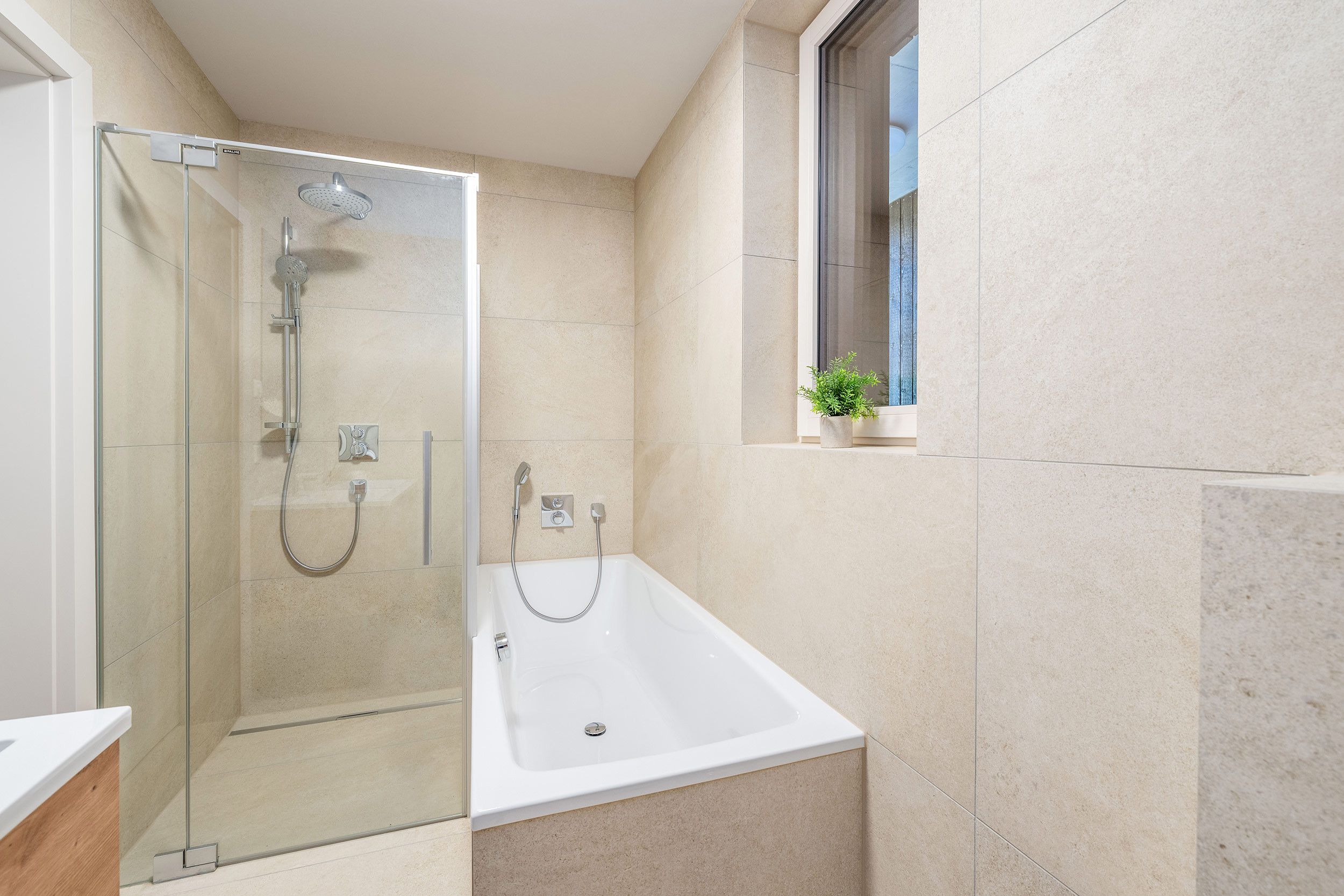 Ein modernes Badezimmer mit Duschkabine aus Glas, einer Badewanne und einem kleinen Fenster mit Pflanze.
