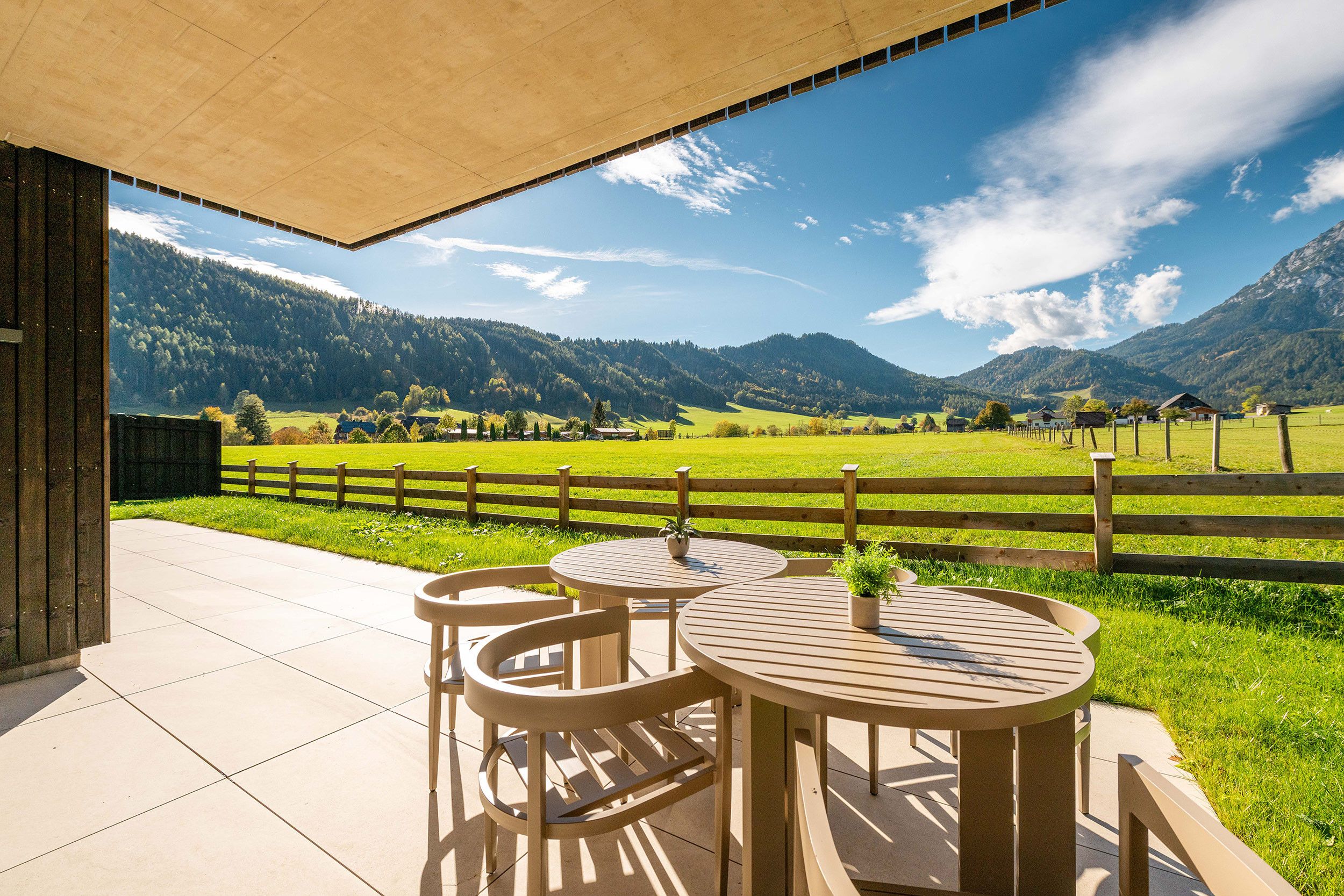 Überdachte Terrasse mit zwei runden Tischen und Stühlen, mit Blick auf ein grünes Feld und Berge.