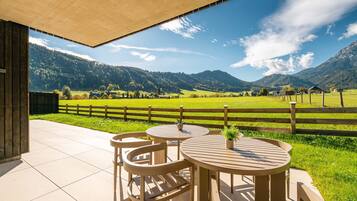 Überdachte Terrasse mit zwei runden Tischen und Stühlen, mit Blick auf ein grünes Feld und Berge.