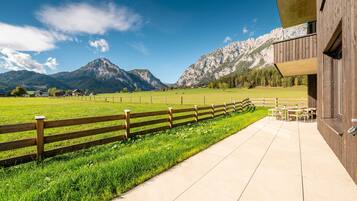 A wooden building with a paved walkway and a wooden fence leading to a green field with mountains in the background.
