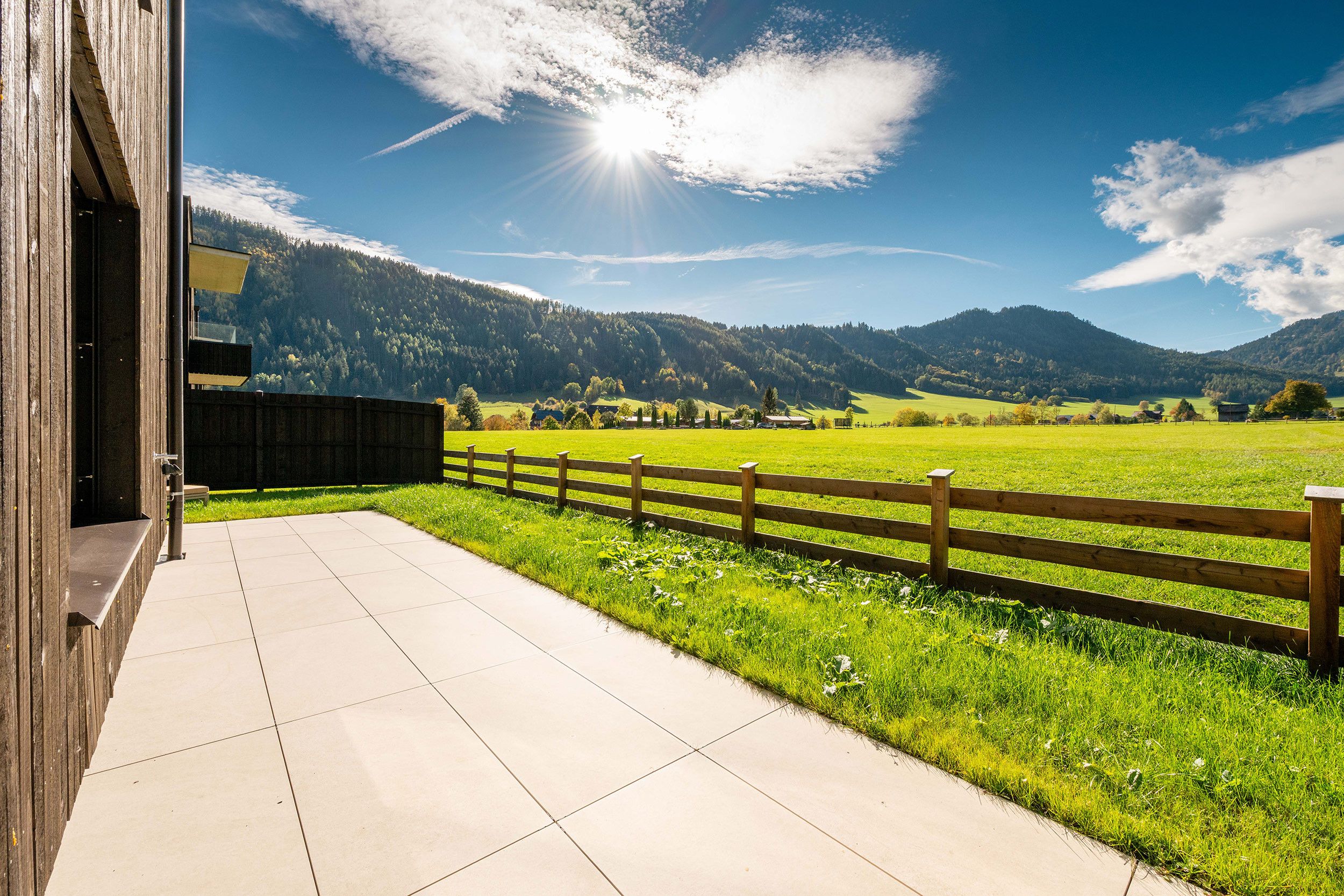A paved pathway leading to a wooden fence overlooking a green field and forested hills.