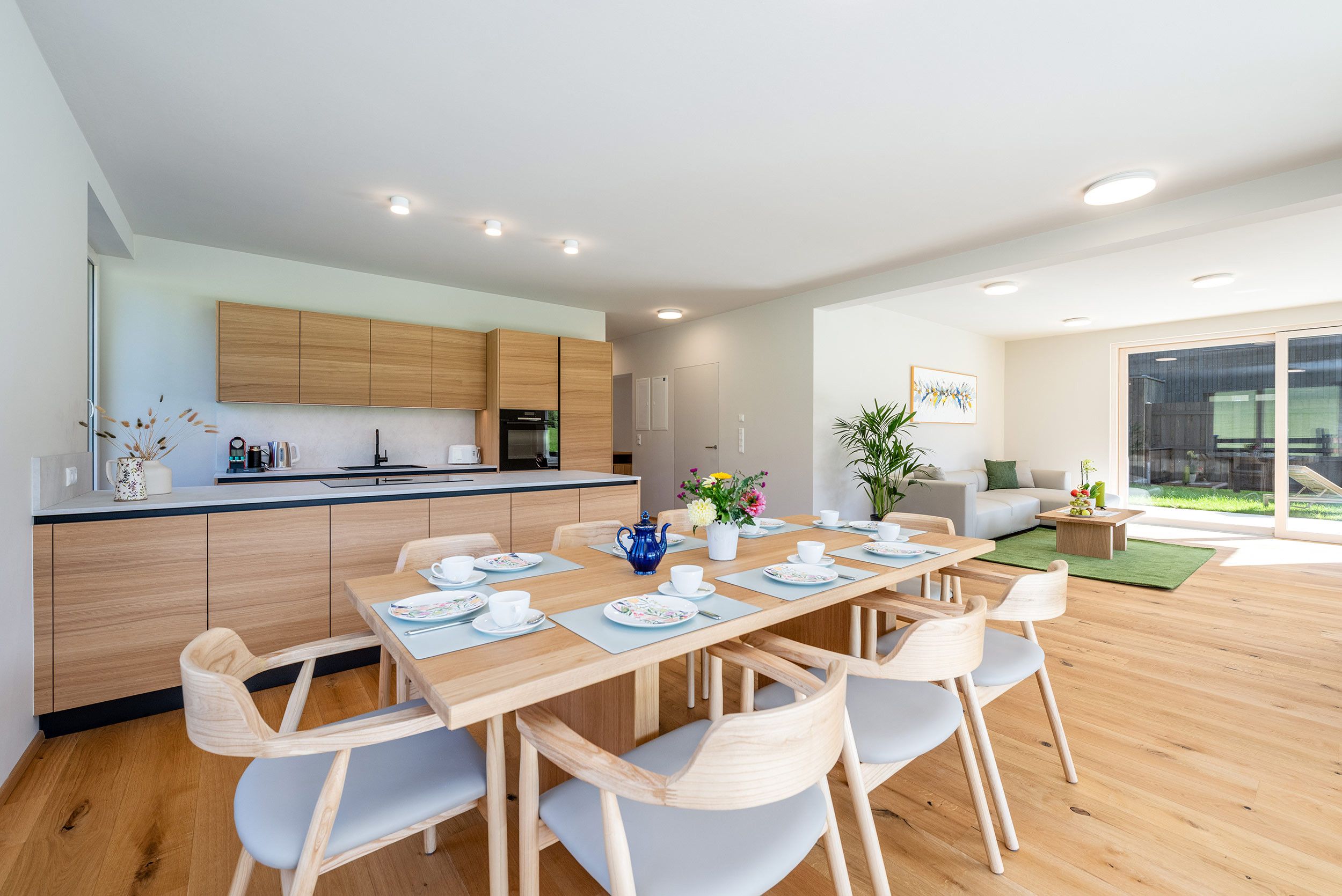Een moderne keuken met houten kastjes, een eethoek met een gedekte tafel voor thee, en een woongedeelte met een bank en een plant.
