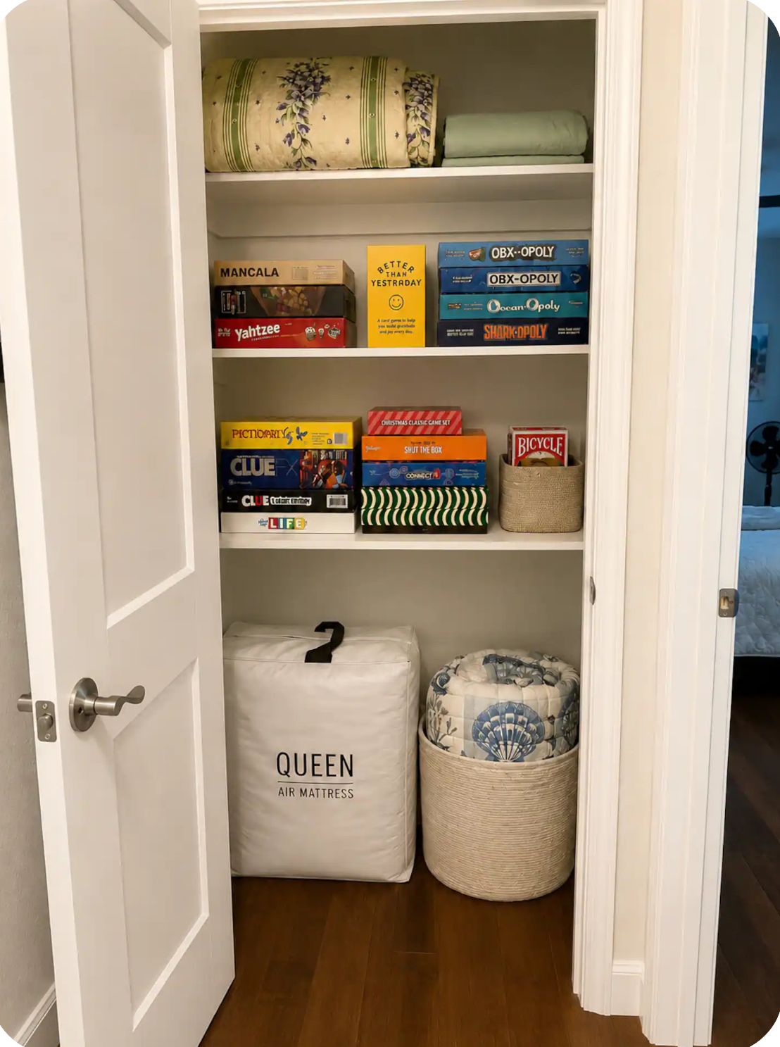 A closet with shelves containing board games, a Queen Air Mattress, and a basket with a patterned cushion.