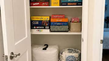 A closet with shelves containing board games, a Queen Air Mattress, and a basket with a patterned cushion.
