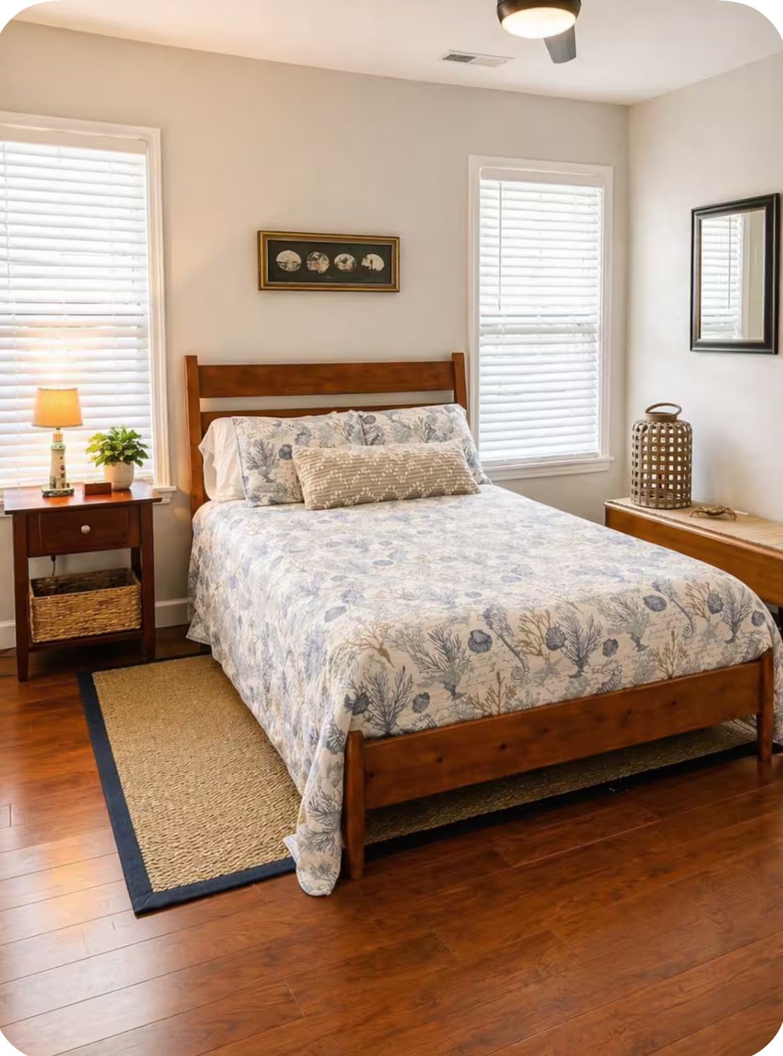 A bedroom with a wooden bed, a bedside table with a lamp, a framed picture on the wall, and a window with blinds.