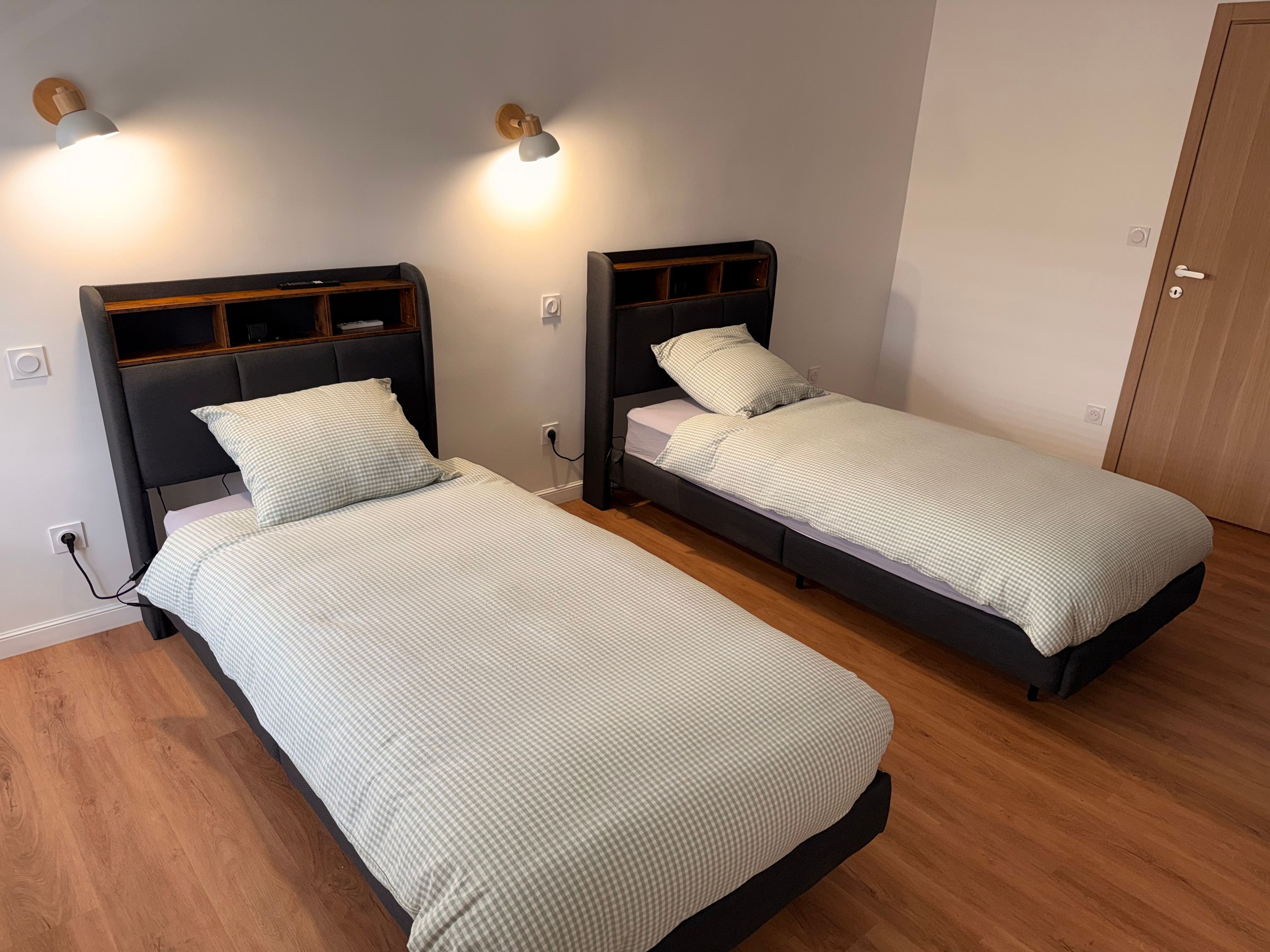 Deux lits simples avec des têtes de lit, un plancher en bois et des lampes fixées au mur.