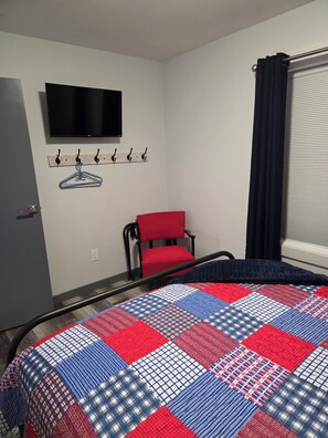 A hotel room with a bed, a television mounted on the wall, a red chair, and a window with curtains. - The Upper Haven - Cozy rural suite (Saint Brieux)