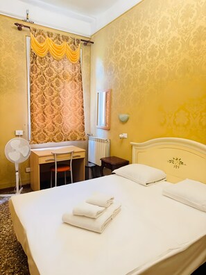 Ein Hotelzimmer mit Bett, Schreibtisch, Stuhl, Ventilator, Spiegel und Heizkörper.