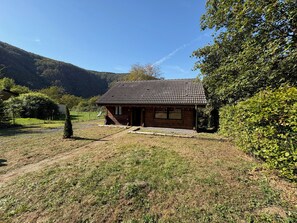 Une cabane en bois avec un toit en tuiles, entourée d’une pelouse et d’un feuillage dense.