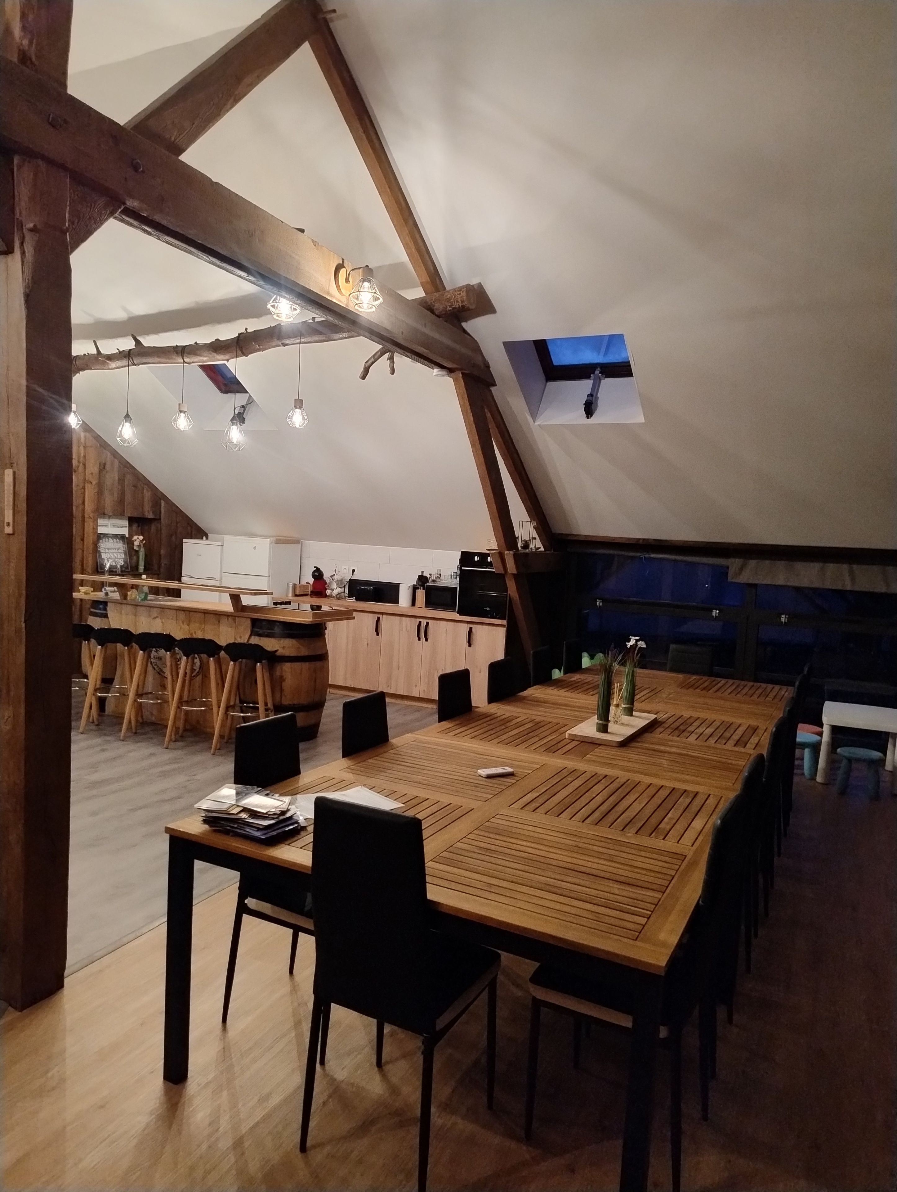 Een ruime eethoek met een lange houten tafel en stoelen, een keuken op de achtergrond, en een schuin dak met zichtbare houten balken.