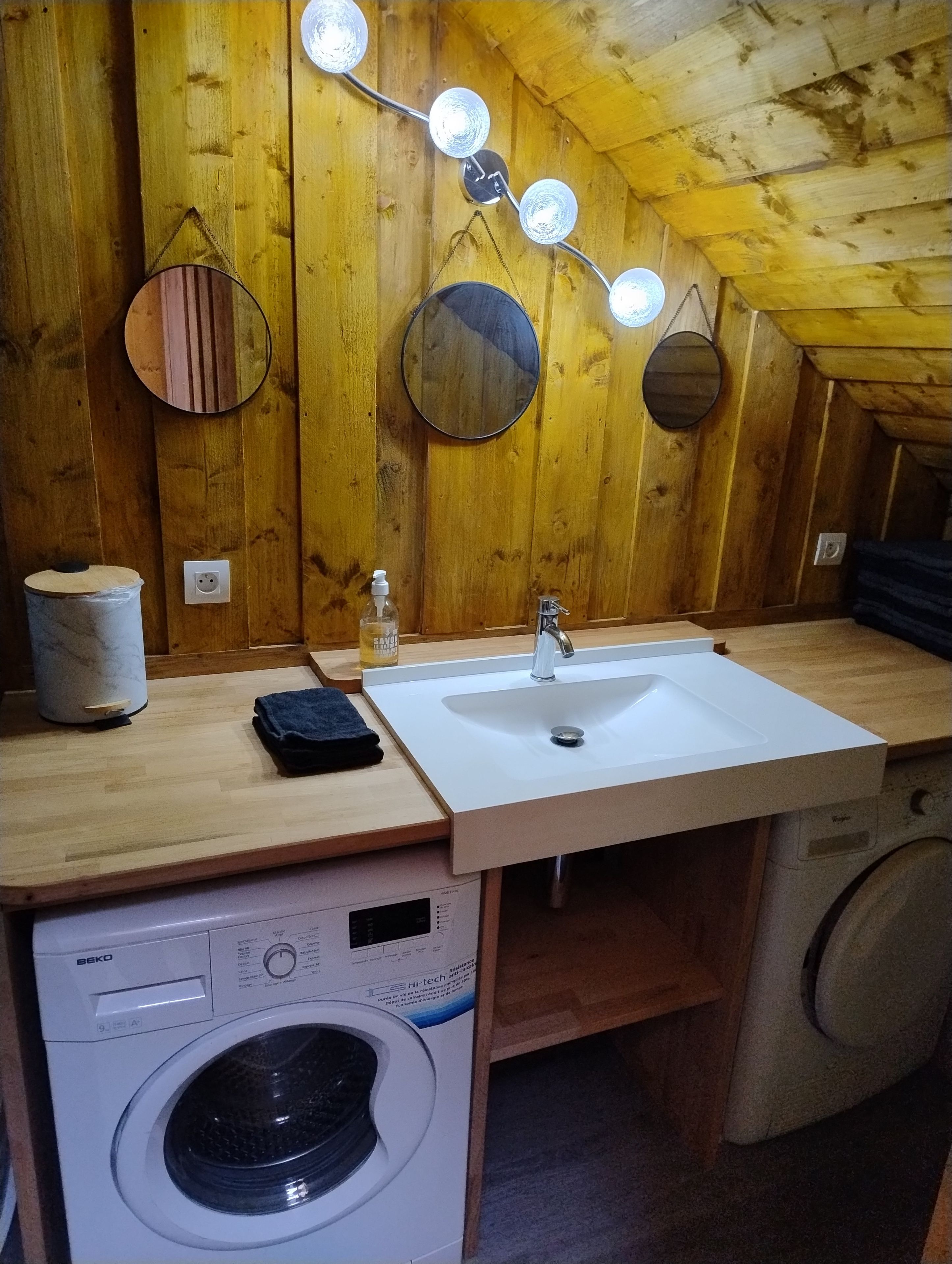 A compact kitchenette with a sink, a washing machine, and a mirror with lights.