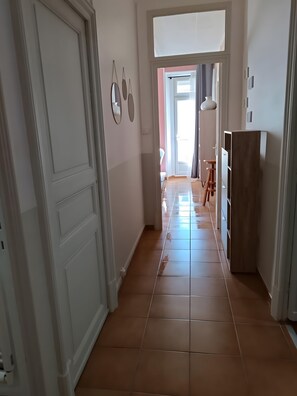 A hallway with a white door, a wooden cabinet, and a tiled floor leading to a room with a window. - Apartment 'Proche Gare' with Balcony, Wi-Fi and Air Conditioning (Lézignan-Corbières)