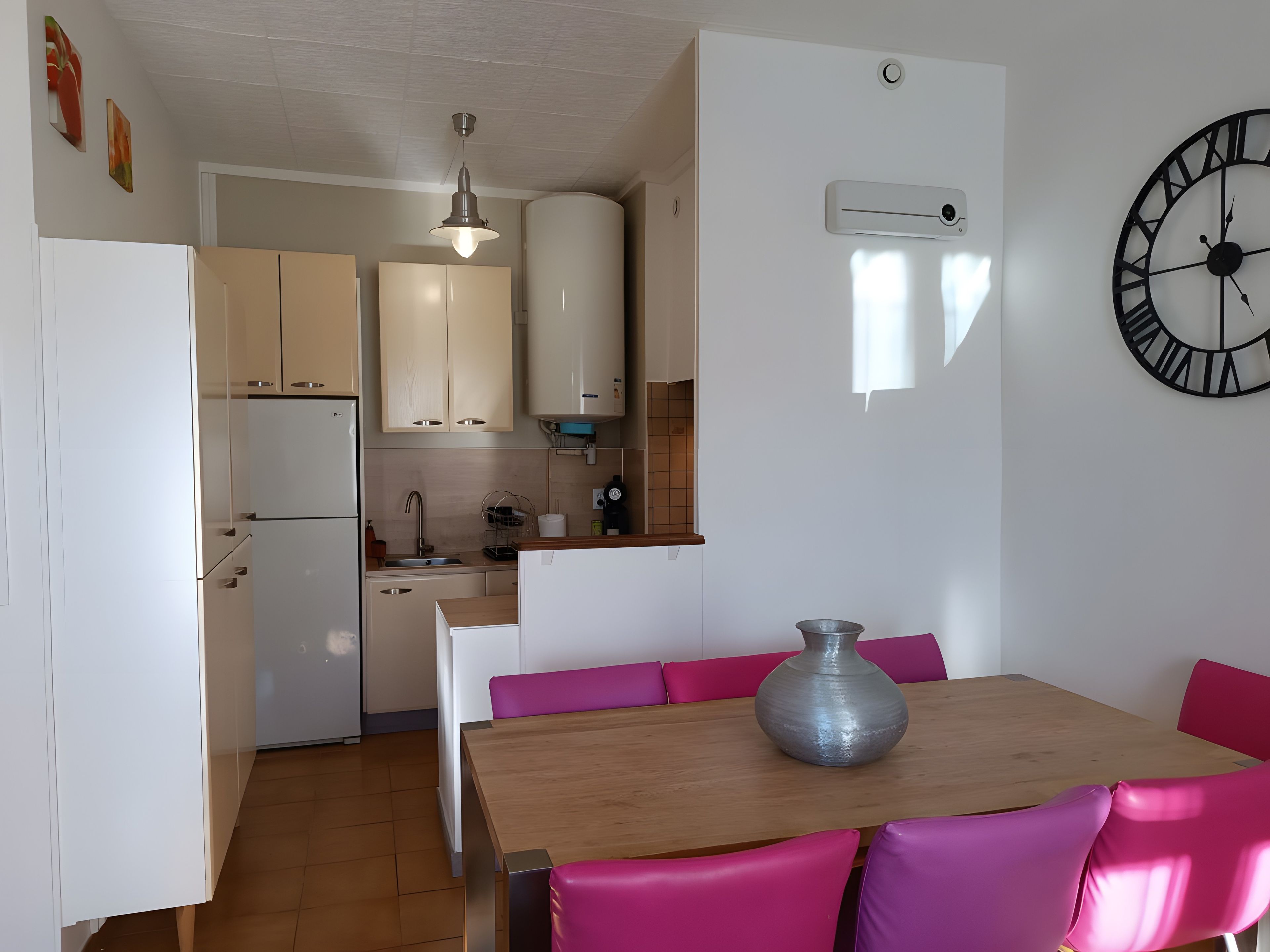 A compact kitchen with a dining area, a clock on the wall, and a vase on the table.