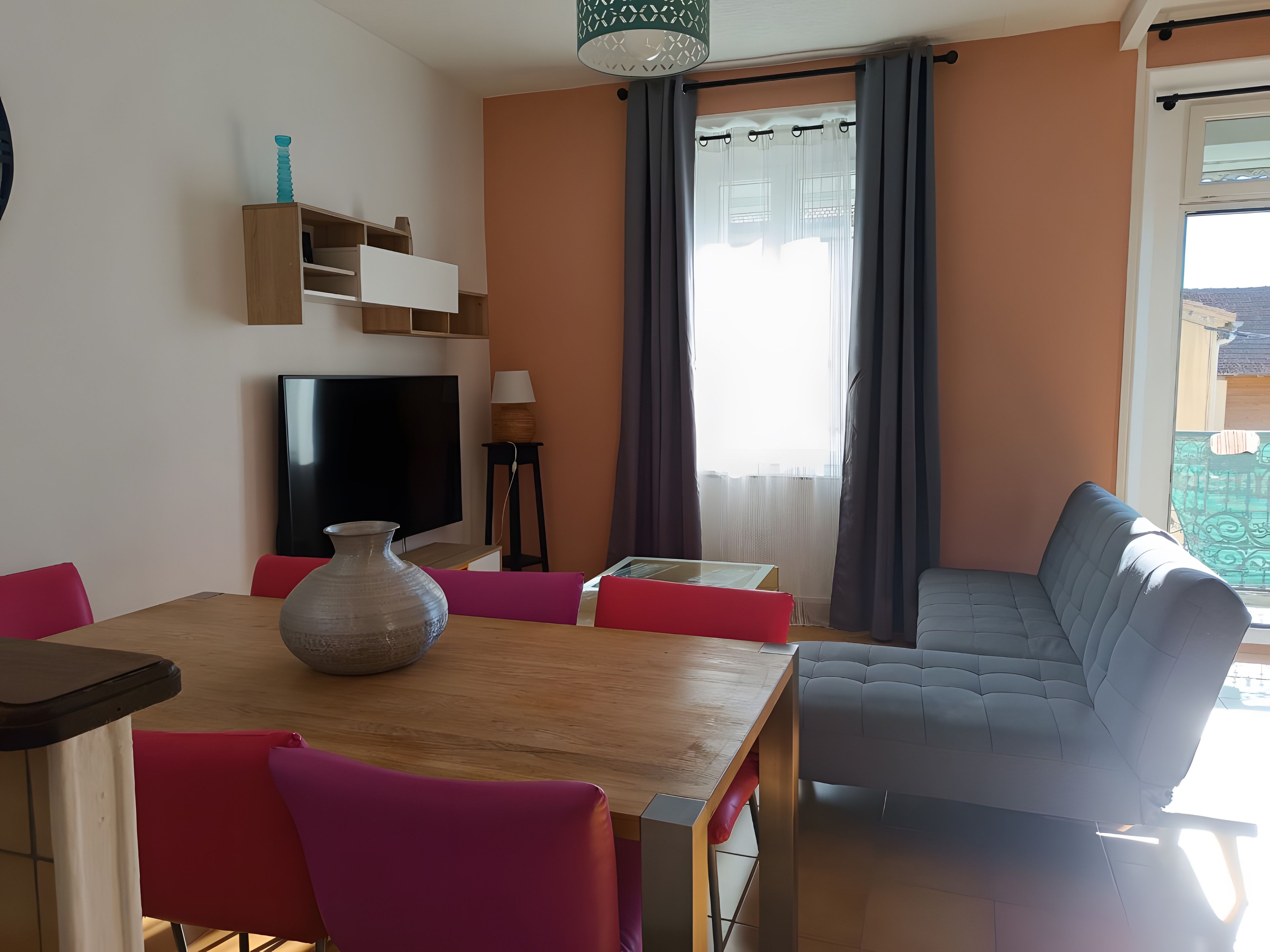 A living room with a sofa, a TV, a table with chairs, a vase, and a window with curtains.