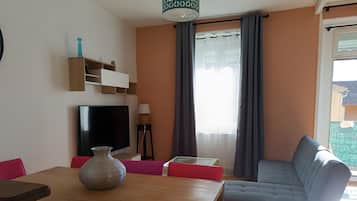 A living room with a sofa, a TV, a table with chairs, a vase, and a window with curtains.