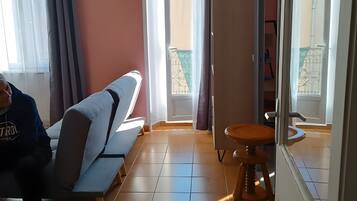 A room with a sofa, a stool, a window with curtains, and a door leading to an outdoor balcony.