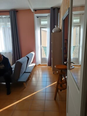 Una habitación con sofá, un taburete, una ventana con cortinas y una puerta que da a un balcón exterior.