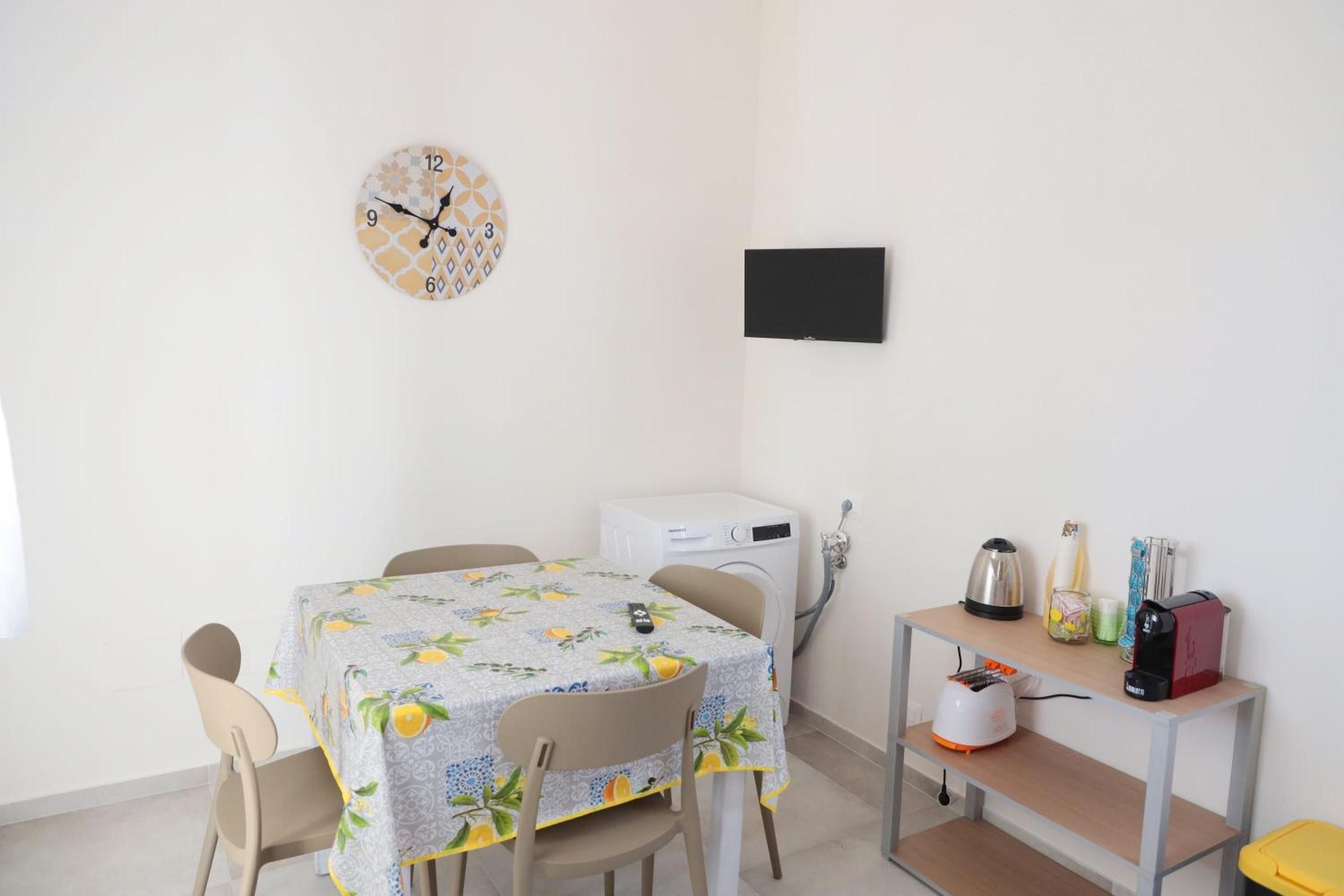 Een kleine, schone kamer met een eettafel, stoelen, een klok, een wasmachine en een klein plankje met diverse spullen.
