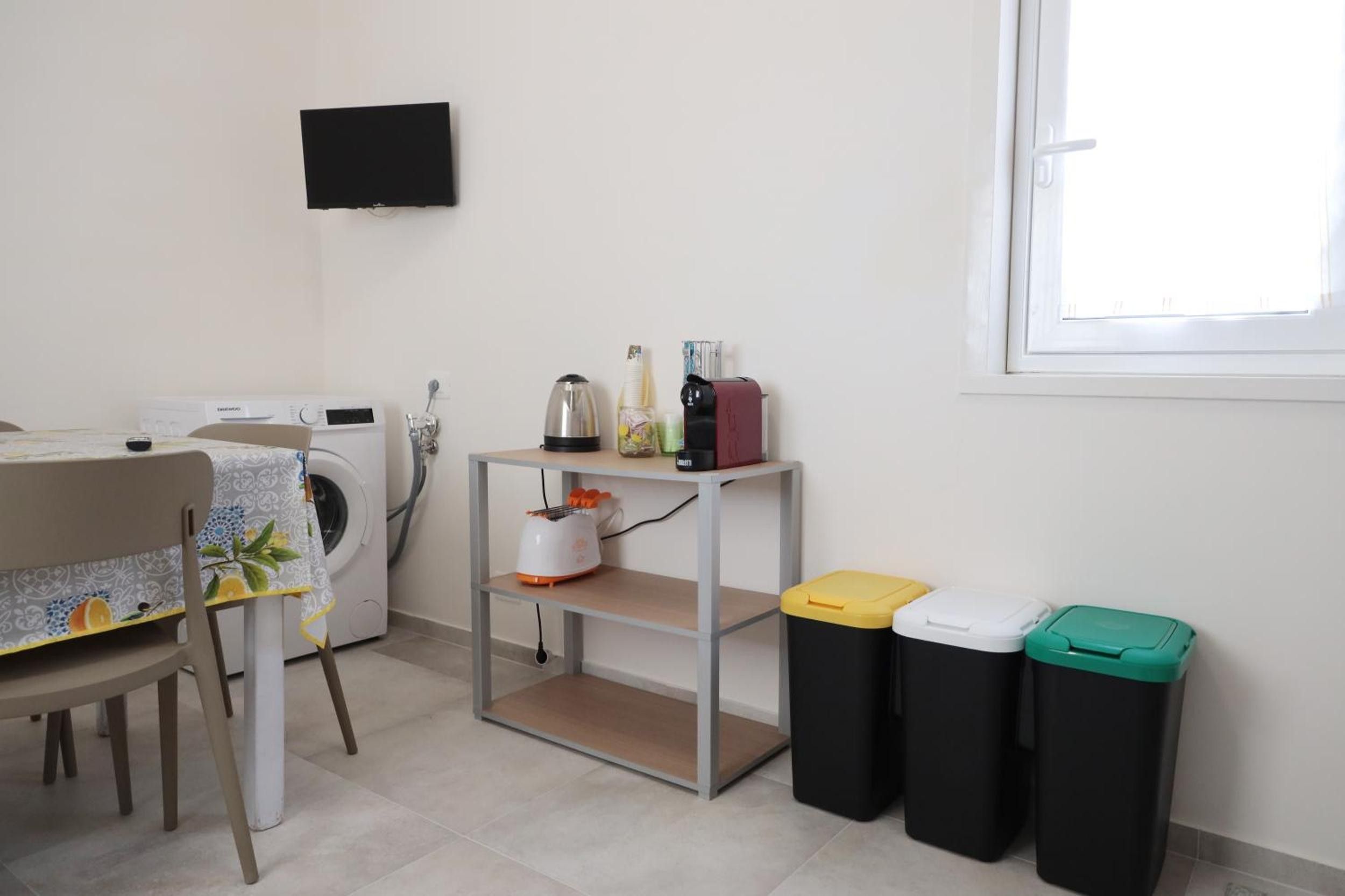 A laundry area with a washing machine, a drying rack, a table with a tablecloth, a shelf with various items, and trash bins.