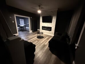 Un salon doté d’une cheminée, d’un téléviseur à écran plat fixé au mur et d’un coin repas visible à travers une porte ouverte.