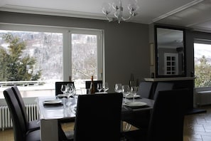 Comedor con mesa lista para cuatro, ventanas amplias y vista a árboles.