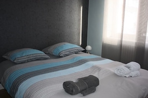 Habitación de hotel con cama, almohadas, una manta, una toalla enrollada y una mesita de noche con lámpara.