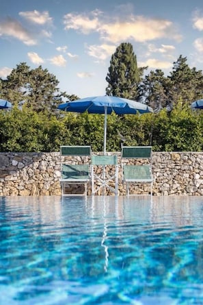 A swimming pool with clear blue water, surrounded by a stone wall and green chairs under blue umbrellas.