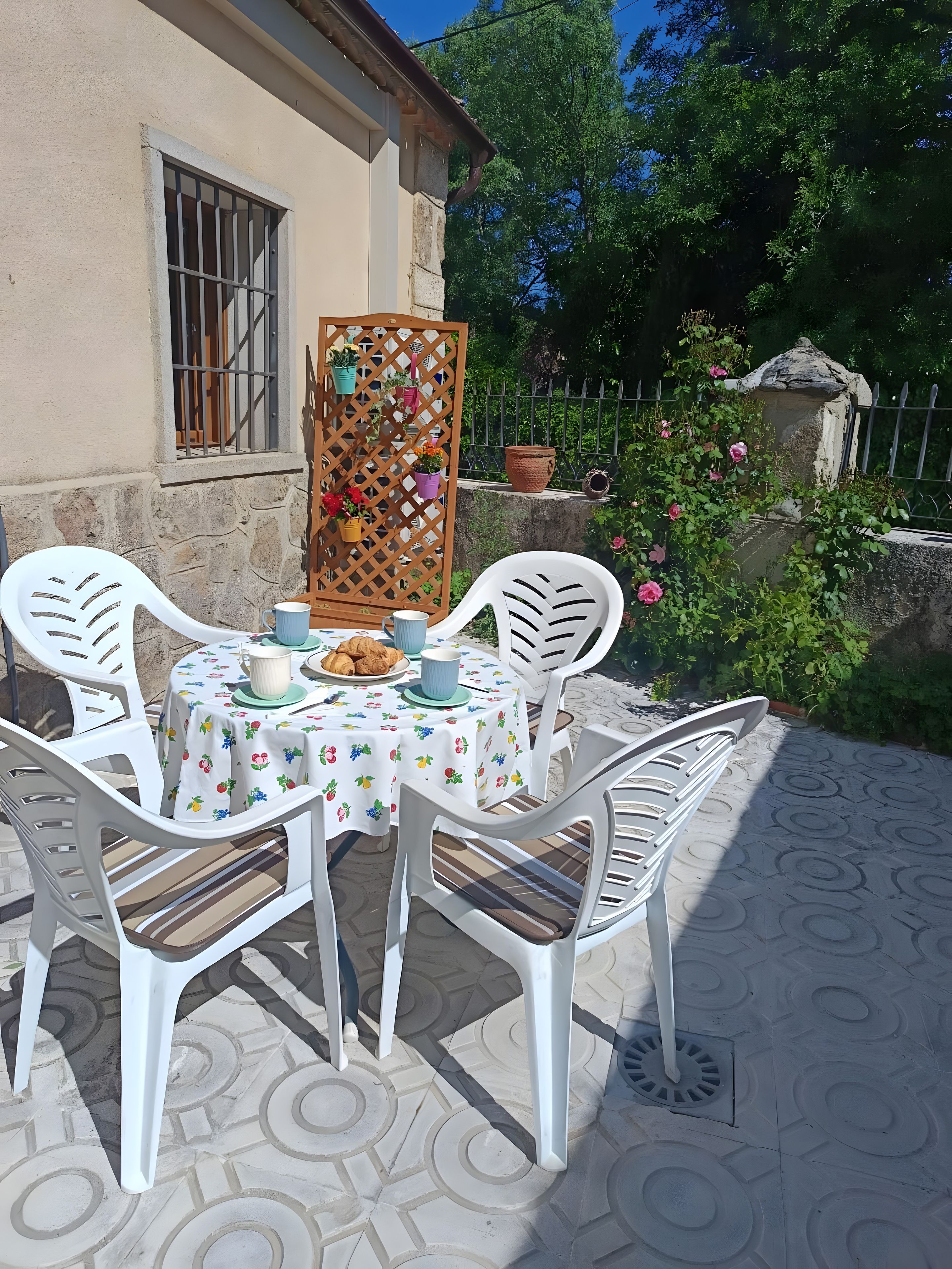 Eine Terrasse mit einem runden Tisch, weißen Plastikstühlen und einer Tischdecke mit Obstmotiven.