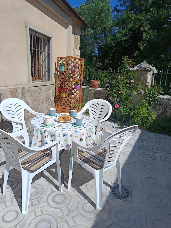 Eine Terrasse mit einem runden Tisch, weißen Plastikstühlen und einer Tischdecke mit Obstmotiven.