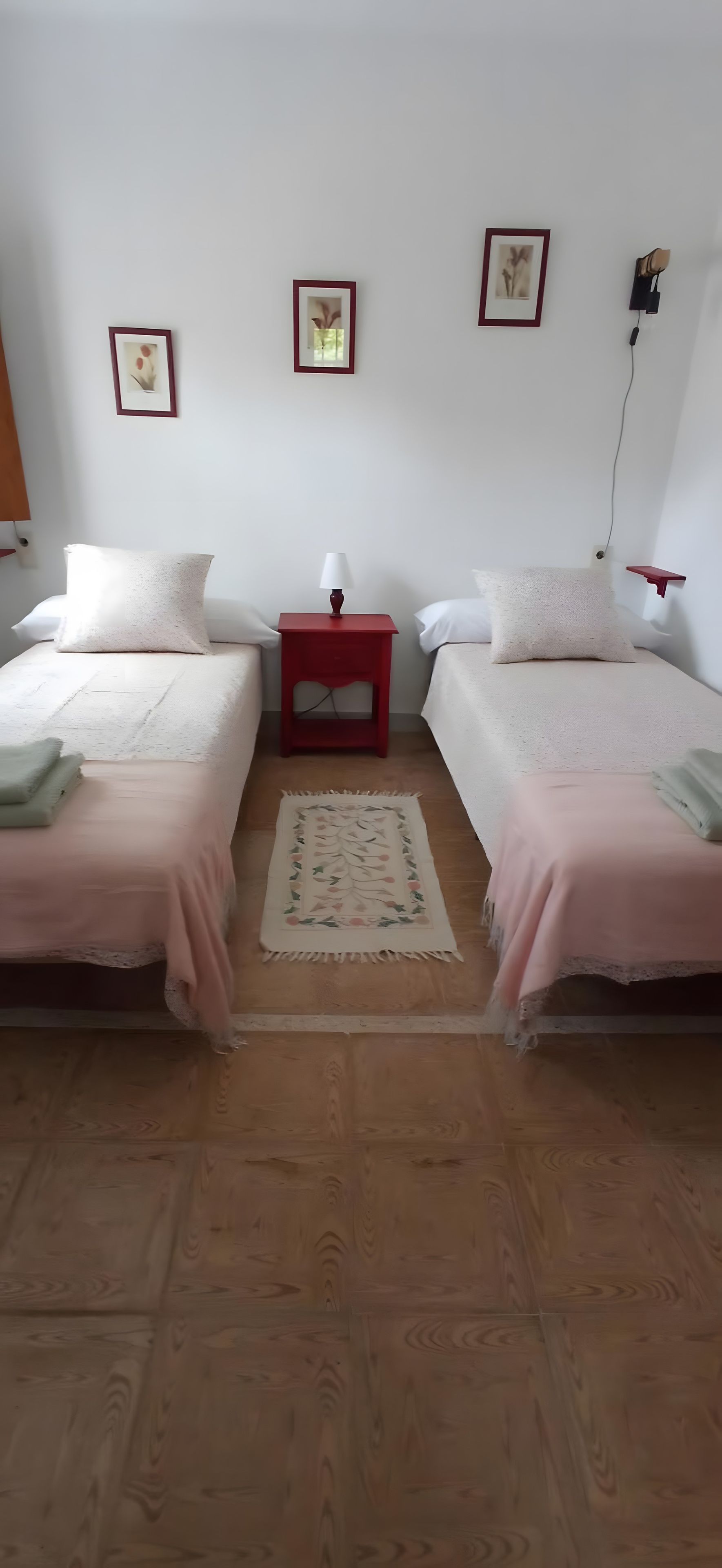 Ein Zimmer mit zwei Betten, einem kleinen Nachttisch mit Lampe, einem Teppich und gerahmten Bildern an der Wand.