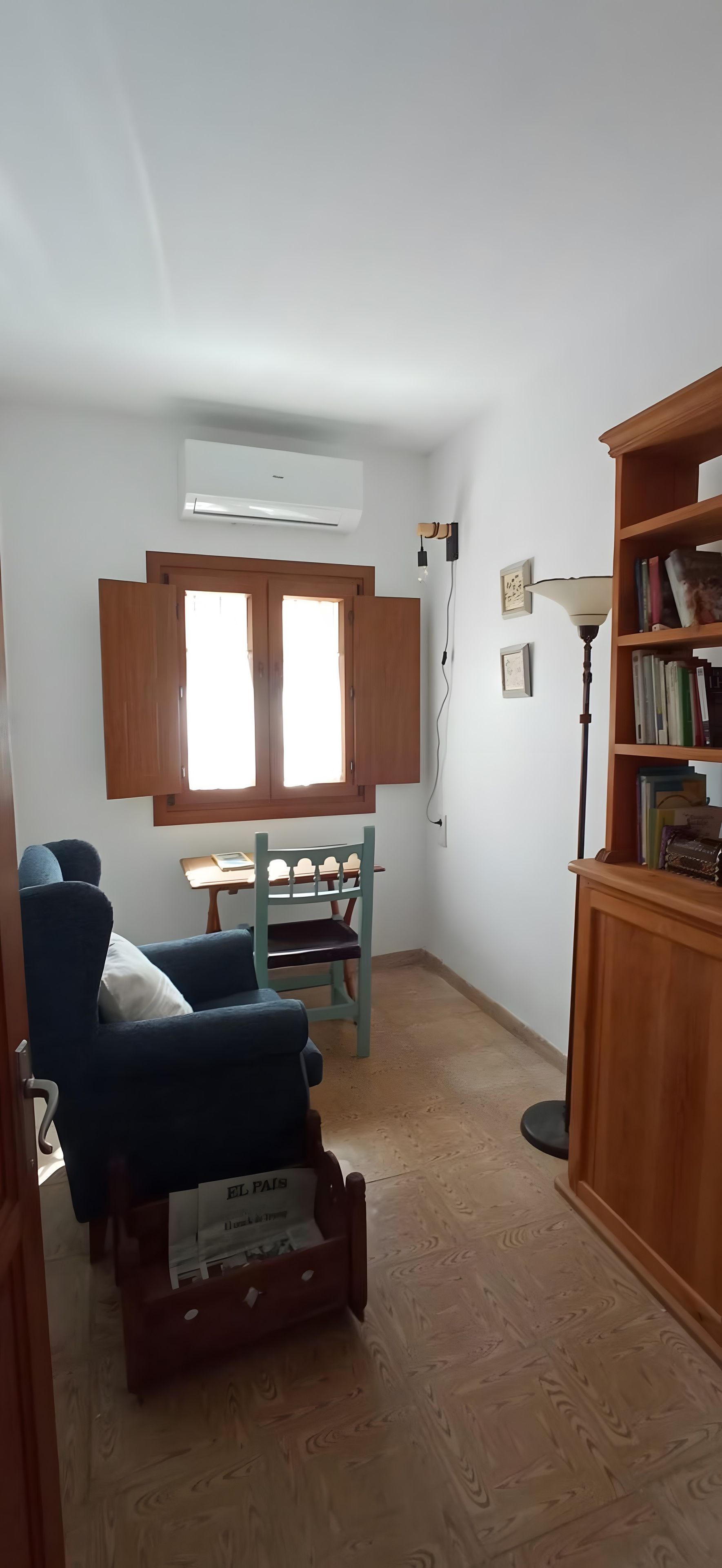 Una habitación con una silla de madera, una mesita, un equipo de aire acondicionado, una estantería y una ventana con contraventanas de madera.