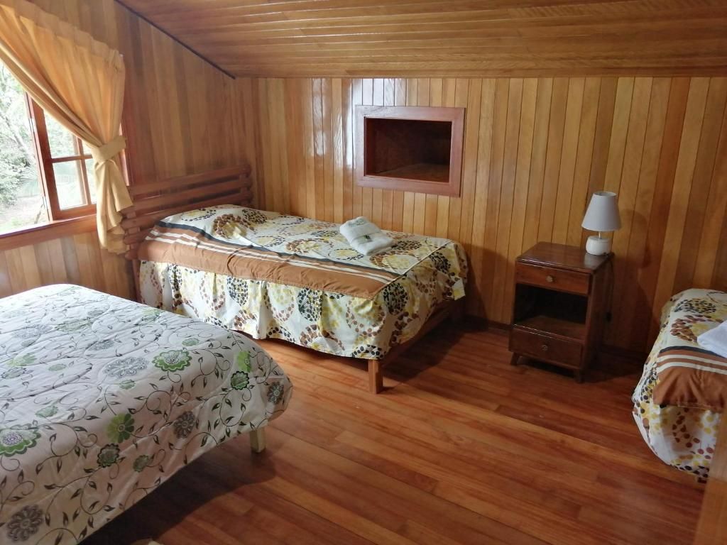 Quarto com duas camas, paredes de madeira, uma pequena mesa de cabeceira com abajur e uma janela com cortinas.